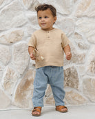 Child wearing a beige shirt and light blue baggy jeans standing against a stone wall.