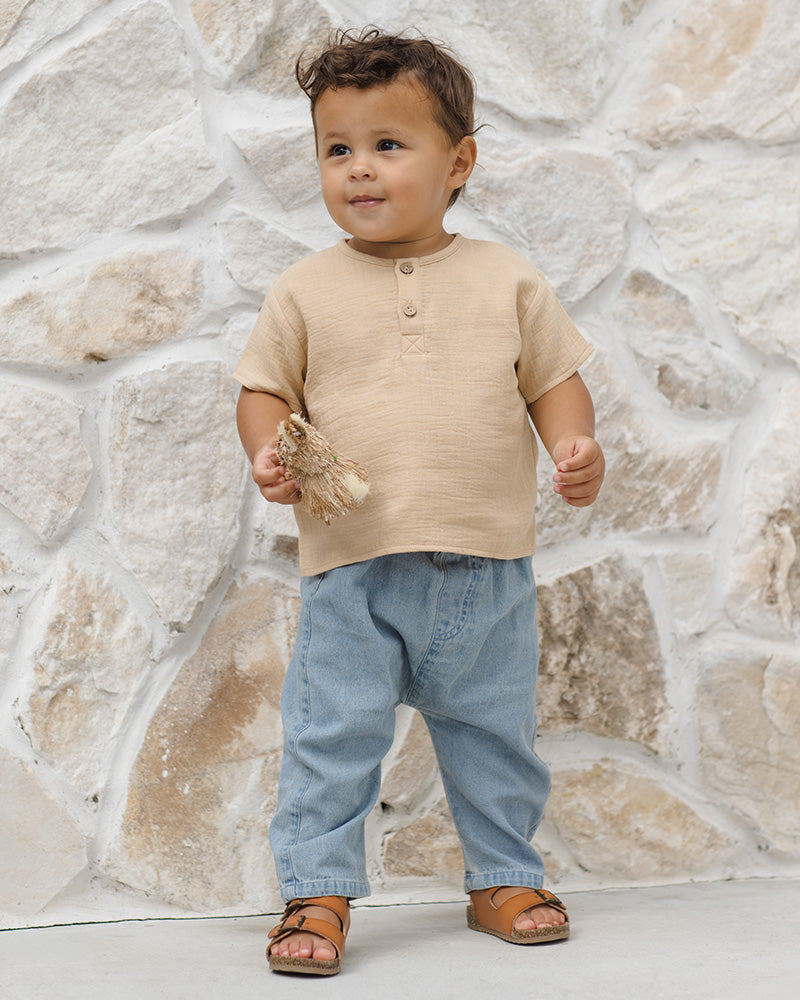 Child wearing a beige shirt and light blue baggy jeans standing against a stone wall.