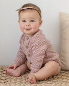 Baby wearing a pink knitted cardigan sitting on a textured surface with a neutral background