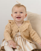 Baby wearing a beige knitted cardigan with a ruffled collar.