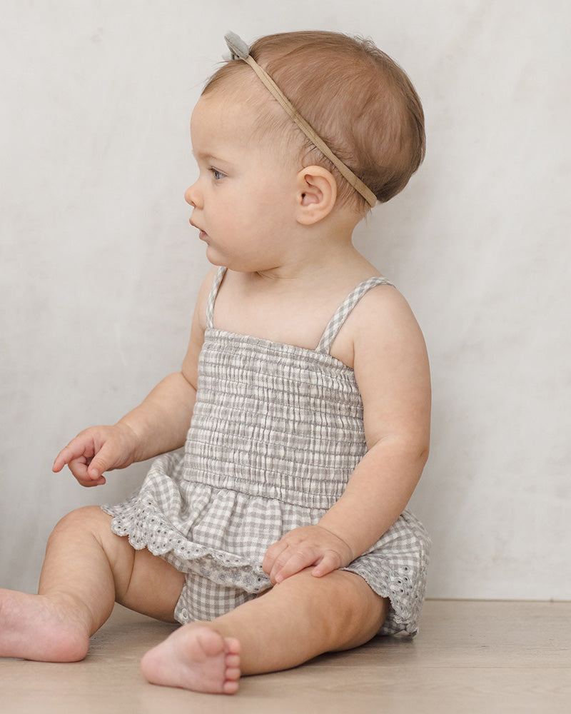 Baby wearing a checkered romper with lace detailing, sitting on a wooden floor against a plain background