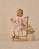Young child in a pink checkered dress sitting on a wooden chair with a vase of flowers on a beige background