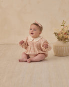 Baby in a pink checkered outfit with a white collar sitting on a beige floor next to a vase of flowers.