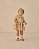 Young child wearing a beige dress with ruffled collar against a beige background