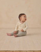Baby sitting on a beige blanket wearing a cream long-sleeve shirt and sage green pants.