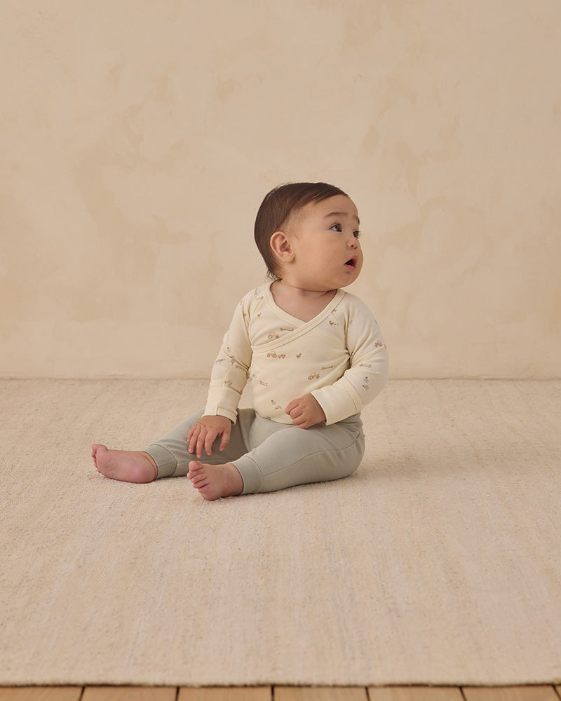 Baby sitting on a beige blanket wearing a cream long-sleeve shirt and sage green pants.