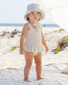 Child in a white striped swimsuit and hat standing on a sandy beach.