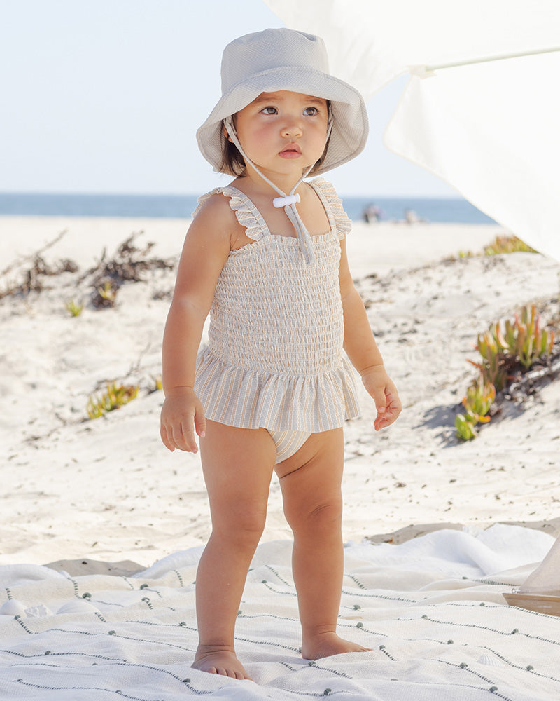 Child in a white striped swimsuit and hat standing on a sandy beach.