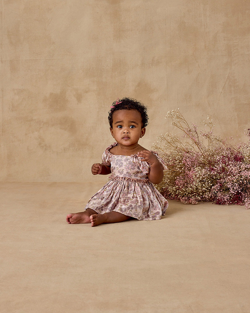 Baby in a floral dress sitting on a beige background with pink flowers.