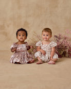 Two young children in floral dresses sitting on a beige surface with flowers in the background.