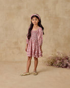 Young girl in a pink floral dress standing against a beige background with dried flowers.