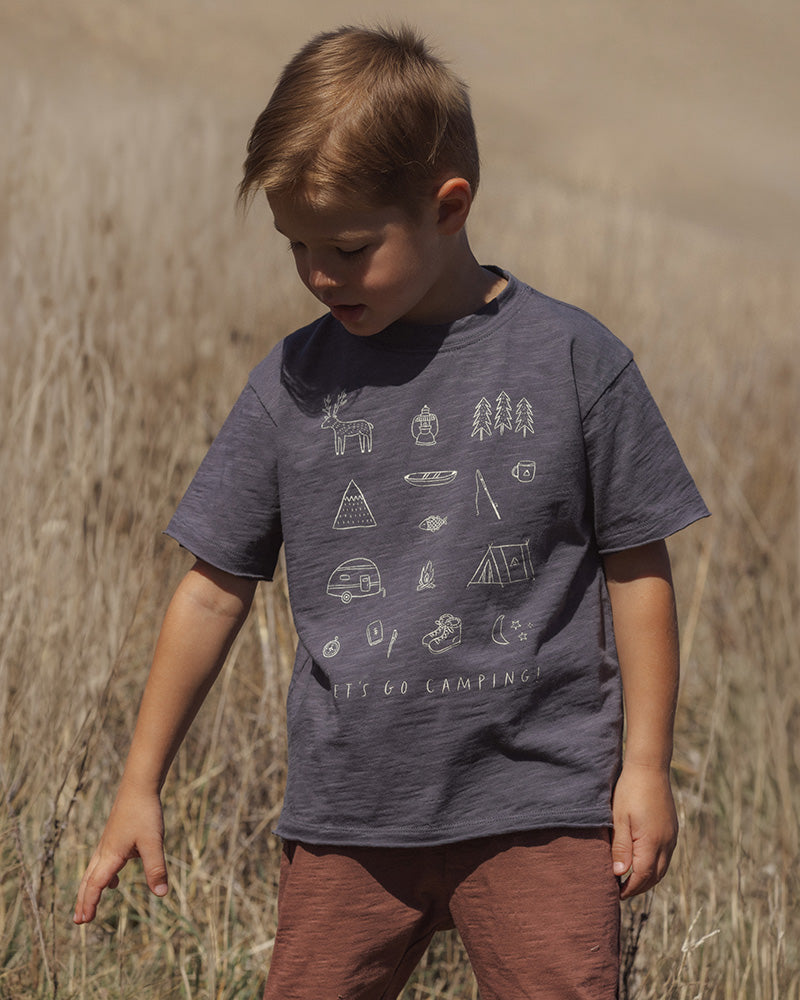 A young boy wearing a dark heather gray t-shirt with white camping-themed graphics and brown pants, standing in a dry grassy field.