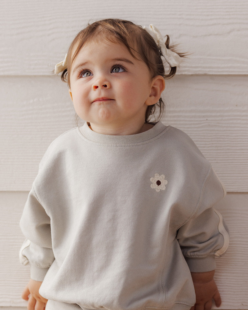 A toddler girl with light brown hair in a white bow, wearing a light gray sweatshirt with a small white flower detail on the chest, leaning against a white paneled wall.