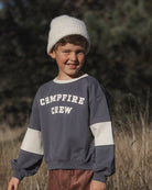 A young boy wearing a gray and white color-block crewneck sweatshirt with 'CAMPFIRE CREW' printed on the chest, paired with a white knit beanie and brown corduroy pants, standing outdoors in a grassy field.