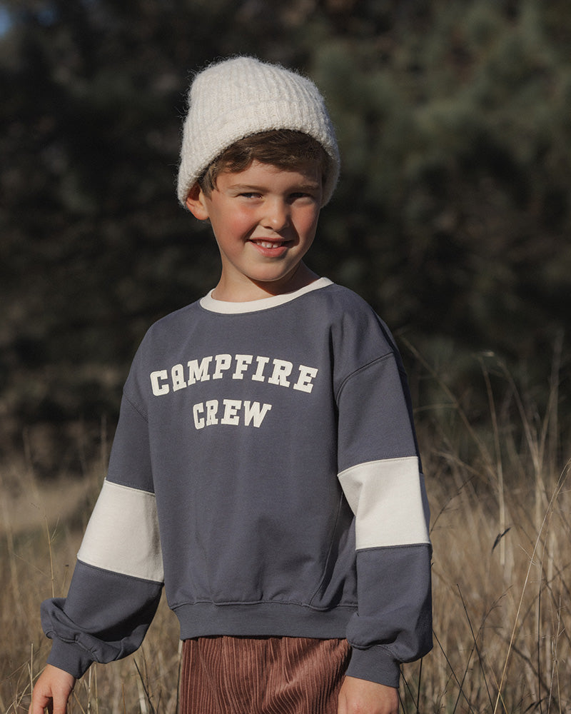 A young boy wearing a gray and white color-block crewneck sweatshirt with 'CAMPFIRE CREW' printed on the chest, paired with a white knit beanie and brown corduroy pants, standing outdoors in a grassy field.