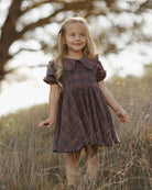 A young girl in a field wearing a dark brown and red plaid dress with a ruffled Peter Pan collar and puff sleeves.
