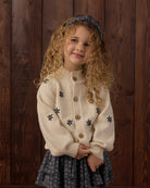A young girl with curly blonde hair wearing a cream-colored knit cardigan with dark blue star motifs standing against a dark wood plank background.