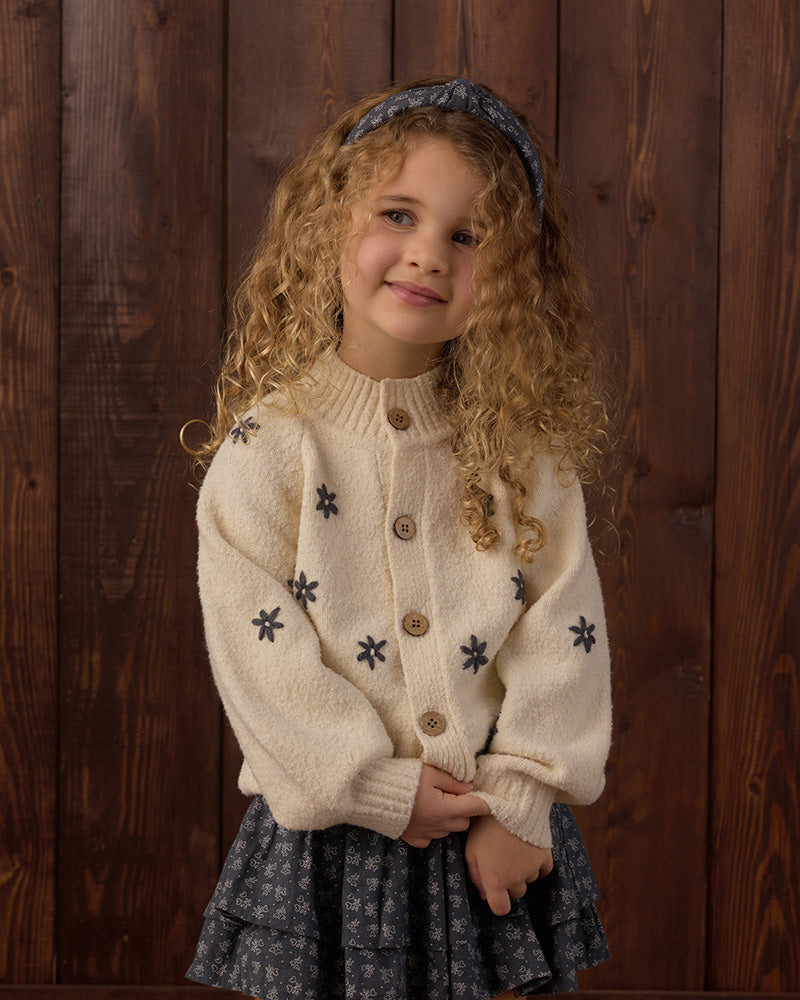 A young girl with curly blonde hair wearing a cream-colored knit cardigan with dark blue star motifs standing against a dark wood plank background.
