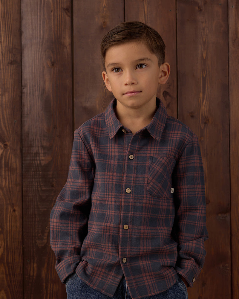 A young boy wearing a long-sleeve button-down plaid shirt in dark blue and red, standing against a dark brown wooden plank wall.