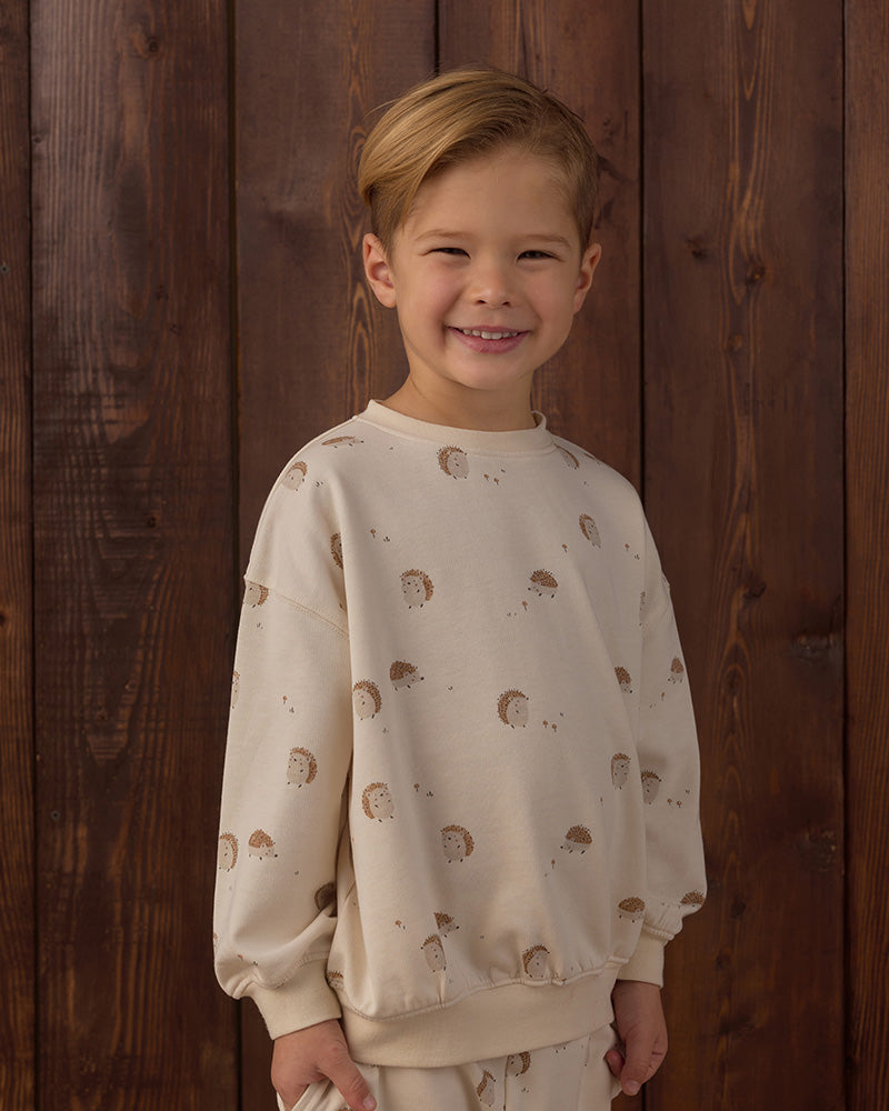 A young boy smiling in front of a dark wood plank wall, wearing a cream-colored sweatshirt with a brown hedgehog pattern and matching sweatpants.
