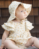 Baby wearing a floral sun hat and romper with ruffle sleeves sitting against a wooden wall.