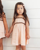 A young girl wearing matching peach dresses with trim against a white wooden background.