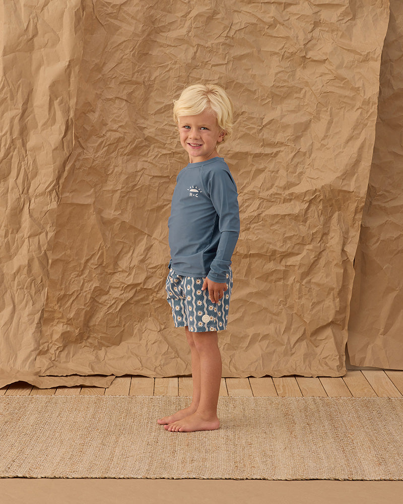 Child wearing a blue shirt and patterned swim shorts standing against a brown paper backdrop