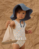 Child wearing a floral swimsuit and blue sun hat holding a toy sailboat against a brown textured background