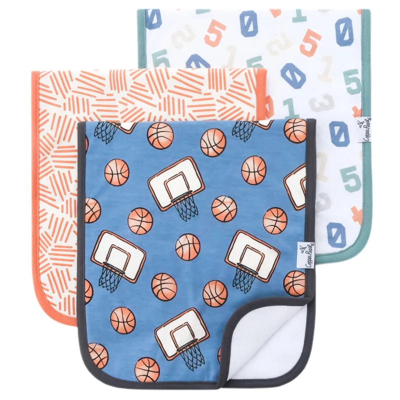 A set of three burp cloths with patterned designs. One has a blue background with basketballs, another has a white with orange numbers, and the last one has a geometric pattern with orange and white.
