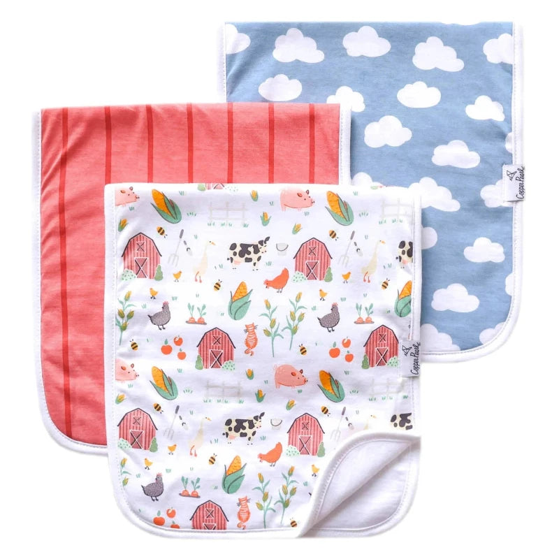 A set of three burp cloths with farmstead designs, including patterns of barns, clouds, and animals on a white background, and a solid blue burp cloth.