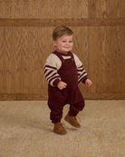 Child wearing a striped sweater and maroon overalls standing against a wooden paneled wall.