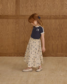 Young girl in a floral skirt and navy top standing against a wooden paneled wall.