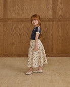 Young girl in a navy top and floral skirt standing against a wooden paneled wall.