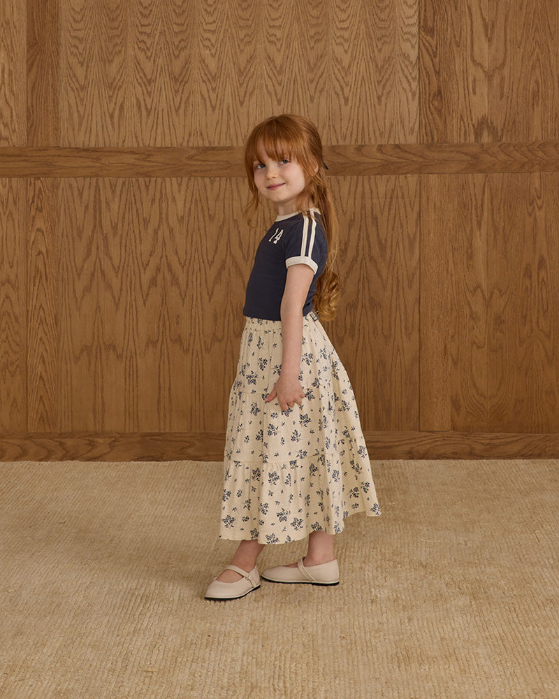 Young girl in a navy top and floral skirt standing against a wooden paneled wall.
